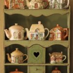 Cottage-shaped ceramic teapots displayed on a rustic green wooden shelf unit.