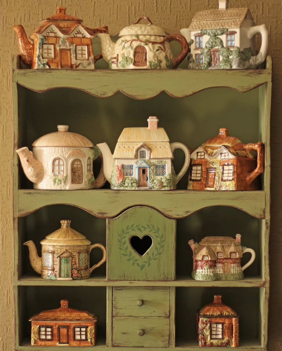 Cottage-shaped ceramic teapots displayed on a rustic green wooden shelf unit.