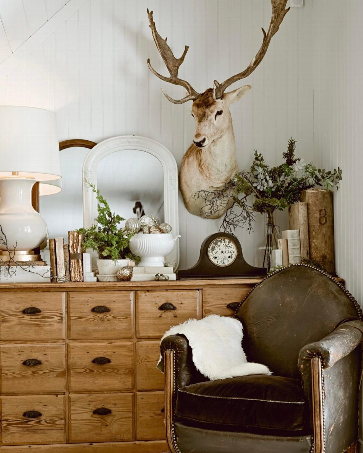 Mounted deer head on a white wall above a rustic wooden dresser and armchair.