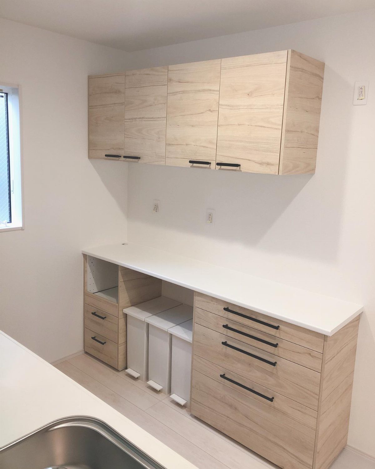 Light wood kitchen cabinets with white countertops and integrated pull-out trash bins.
