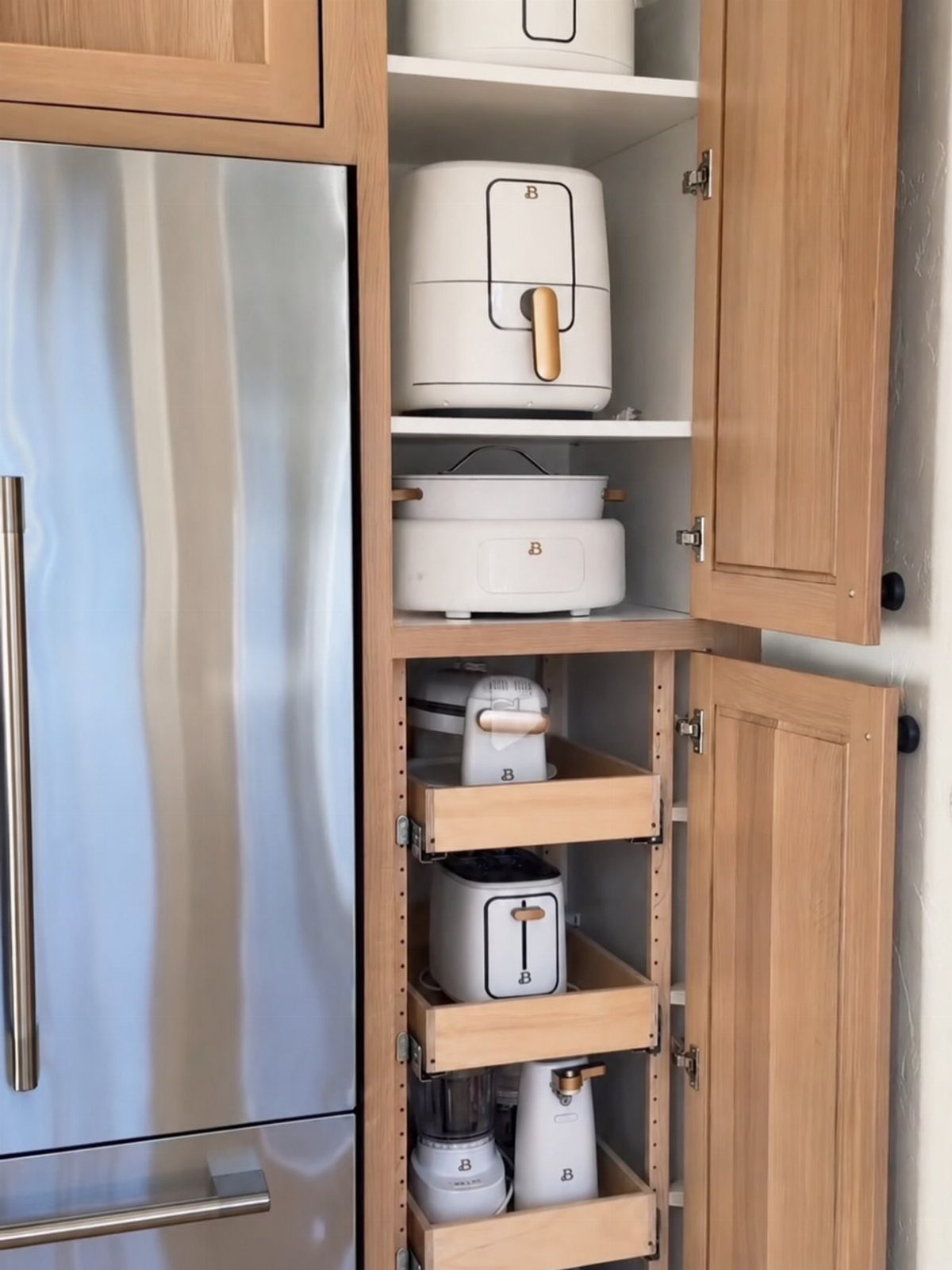Wooden kitchen cabinet with pull-out shelving holding white appliances next to a refrigerator