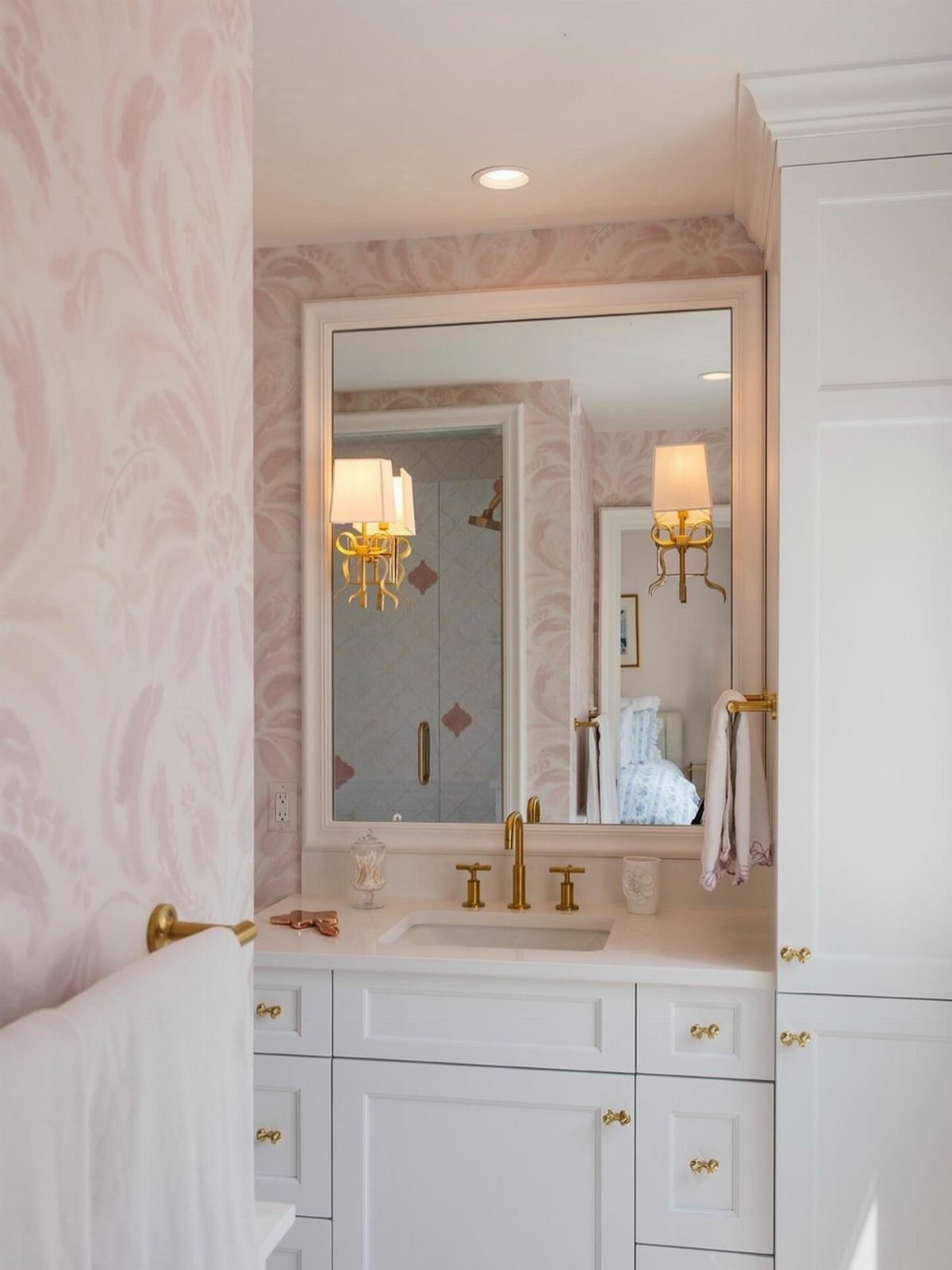 White bathroom vanity with gold fixtures and pink patterned wallpaper in a bright room