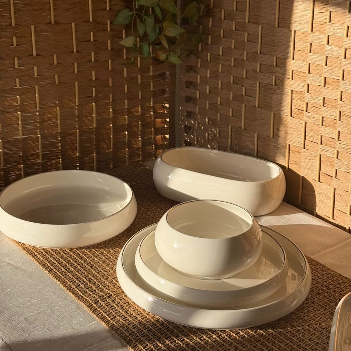 White stoneware dishes including bowls and plates on a woven mat by a bamboo wall.